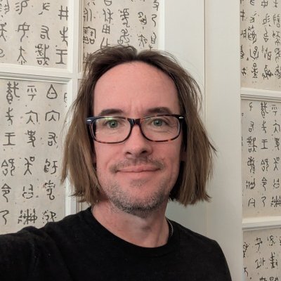 Portrait of Cody Collier, a man with brown hair and glasses, standing in front of a background featuring calligraphic art.